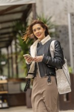 Happy businesswoman using mobile phone while walking down the street, carrying a shopping bag