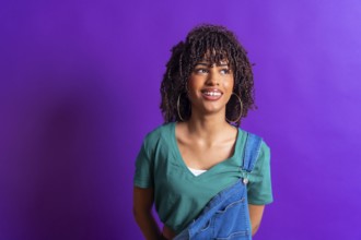 Young woman with curly hair, wearing a green t shirt and denim overalls, smiles while looking up