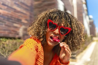 Cool latin young woman wearing heart shaped sunglasses taking selfie and eating lollipop