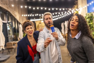 Man and two multiracial women singing karaoke having fun at night party in a terrace of a bar