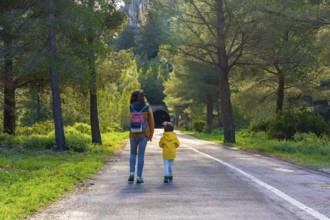 Mother and child enjoying a leisurely walk on a paved road amidst lush greenery and pine trees near
