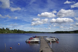 Lake with a wooden jetty and boats moored on it under a blue sky with white clouds, Stora Le,