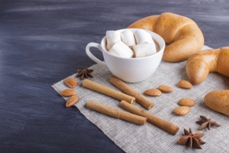 A cup of hot chocolate with marshmallow, croissants and spices on linen napkin and black wooden