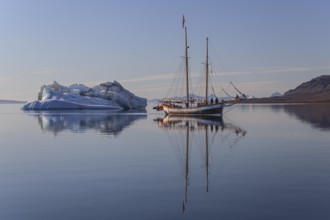 Traditional sailing ship reflected in fjord next to iceberg, sunny, Ittoqqortoormiit, Scoresby