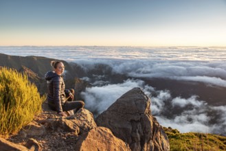 A woman sits on a rocky mountainside and calmly enjoys the wide view over the sea of clouds, Pico