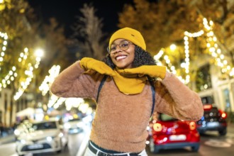 Smiling young woman wearing yellow winter clothes enjoying christmas lights in a city street at