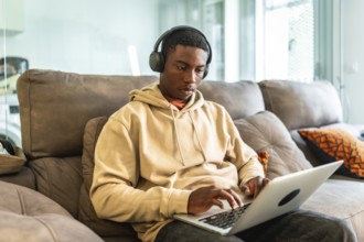 Young black man using laptop and headphones while working or studying from home, sitting on a