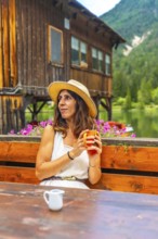 Woman wearing straw hat holding a mug and enjoying a hot beverage at a wooden table with lake