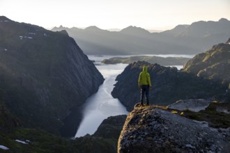 Mountaineer, young man standing on rocks and looking into the distance, at sunrise, fjord