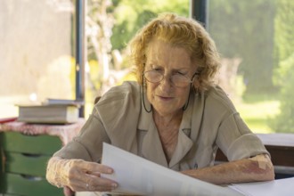 Focused elderly woman wearing glasses reading important documents while sitting at her home office