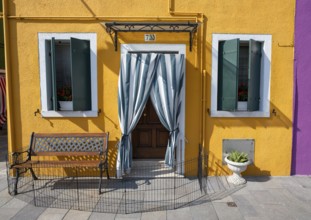 Yellow house facade with entrance door with green and white curtain and windows, colourful houses