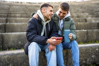 Two men are sitting on steps, enjoying a reusable coffee cup and sharing a laugh as they look at a