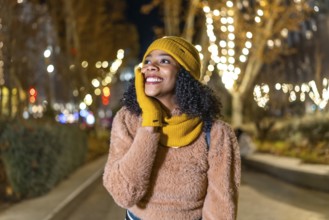 Smiling young woman wearing winter clothes touching her face while enjoying christmas lights in a