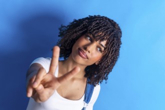 Young woman with curly hair making a victory sign with her fingers against a vibrant blue