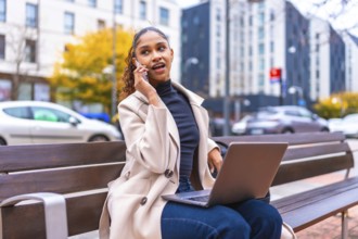 Busy young african american businesswoman using laptop talking to phone sitting on bench in the