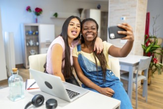 Two happy multi ethnic female students taking a selfie with a smartphone while studying at home