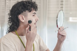 Young man with curly hair and piercings applying black makeup with a brush while holding a mirror,