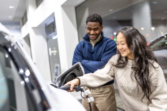 Happy multiethnic couple charging their new electric car at a dealership showroom, embracing a
