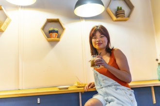 Low angle view portrait of an asian cool young woman smiling at camera sitting drinking coffee in a