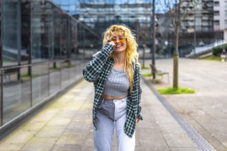 Stylish young woman with blond curly hair and sunglasses dances in a modern urban setting, enjoying