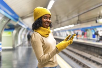 Young black woman wearing yellow winter clothes and gloves using her smartphone while waiting for