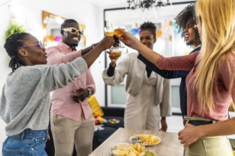Multi-ethnic friends toasting with beer standing around table with snacks in the living room during