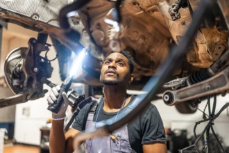 Young mechanic using flashlight, inspecting underside of car in a professional workshop, focused on