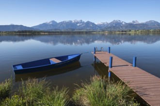 A lake with calm water and blue sky, a boat and a jetty in front of an alpine backdrop, Hopfen am