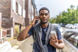 Young african man talking to the mobile commuting to work carrying a hot coffee thermos along a