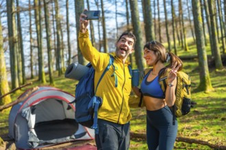 Happy couple of hikers taking a selfie with smartphone in a forest next to their tent during a