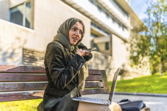 Arab young businesswoman sending voice message with mobile working with laptop sitting on a bench