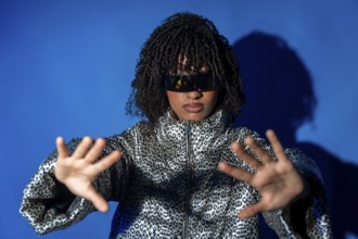 Young woman wearing futuristic visor and patterned jacket, gesturing with hands on blue background,