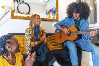 Cool latin man playing guitar sitting on the sofa and enjoying with multi-ethnic female friends at