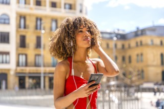 Hispanic woman with curly hair listening to music using mobile at the city