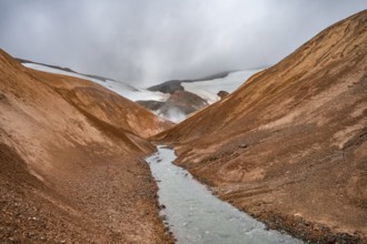 Steaming stream between colourful rhyolite mountains and snowfields, Hveradalir geothermal area,