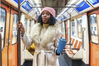 Stylish young black woman wearing winter coat and pink beanie holding reusable coffee cup while