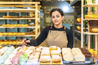 Latin young saleswoman next to a counter full of colorful pastries to sell in a bakery