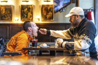 Man feeds his disabled friend in a restaurant, demonstrating care, friendship, and inclusion