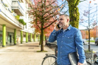 Mature man in denim clothes talking to the mobile and carrying laptop in the city in autumn