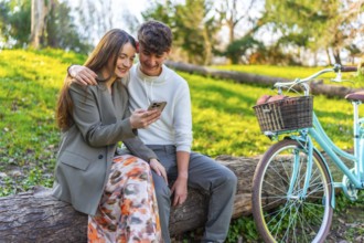 Smiling young couple sitting on a log in a park, sharing a moment on a smartphone, with a vintage