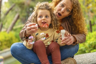 Happy playful mother and girl blowing soap bubbles sitting on a park