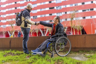 Tourist with backpack shaking hands with a disabled man in a wheelchair in a city park, with a red