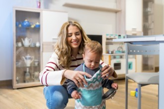 Happy mother helping her cute baby boy wearing a toy apron in the kitchen, playing and having fun