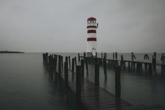 A lighthouse at the end of a wooden jetty under a grey sky over calm water, Podersdorf, Burgenland,