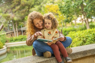 School girl sitting on the lap of her mother reading a book in a park