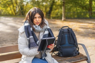 Woman with warm clothes working using digital tablet sitting on a park bench during sunny day of