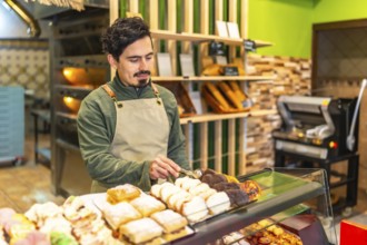 Latin young man arranging sweet artisan pastries on the counter of a bakery shop