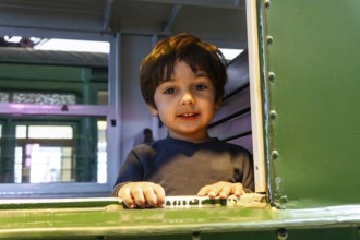 A young boy is sitting in a train car with his hands on the window. He is smiling and looking out