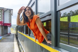 A woman in an orange shirt is looking out the window of a train. She is smiling and waving at the
