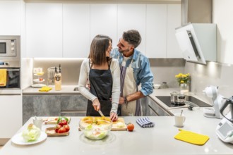 Young couple wearing aprons preparing a vegetable salad together in their modern kitchen, embracing